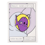 A purple, magic Hanklerfish pin on a grey backing card. The pin is a yellow circle with a purple hanklerfish inside with small hearts on its side. From Karen Hallion.