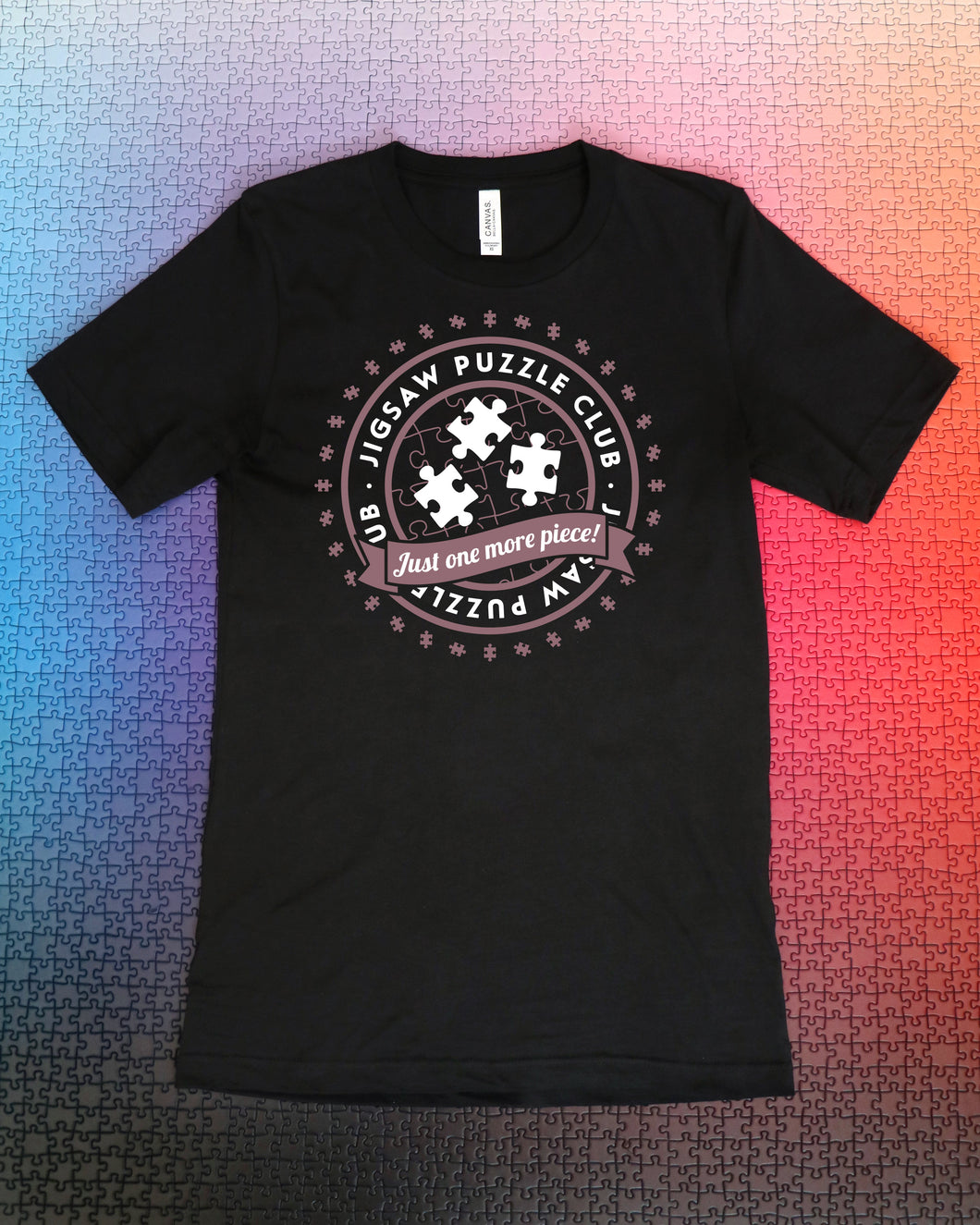 Black t-shirt with the words "Jigsaw Puzzle Club" written in a circle above puzzle pieces and the words "Just One More Piece!" written in a banner. The shirt is laying on top of a gradient puzzle. From Karen Puzzles. 