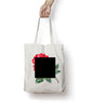 A white tote bag with a red rose with a green stem and leaves covered by a black square - from The Art Assignment