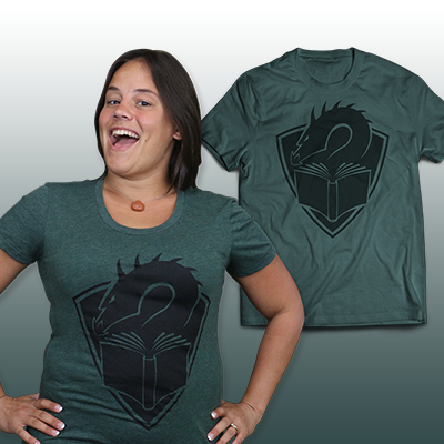 A tealish green colored t-shirt with a centered large black illustration of a dragon's head peering at the pages of an open book, with a crest shape in the background. In front of the shirt is a person with short, black hair wearing the shirt, hands on hips.