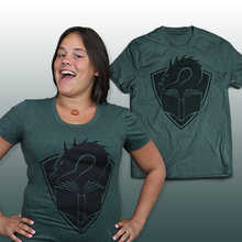A tealish green colored t-shirt with a centered large black illustration of a dragon's head peering at the pages of an open book, with a crest shape in the background. In front of the shirt is a person with short, black hair wearing the shirt, hands on hips.