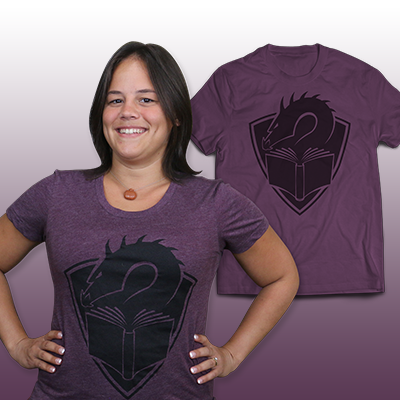 A purple t-shirt with a centered large black illustration of a dragon's head peering at the pages of an open book, with a crest shape in the background. In front of the shirt is a person with short, black hair wearing the shirt, hands on hips.