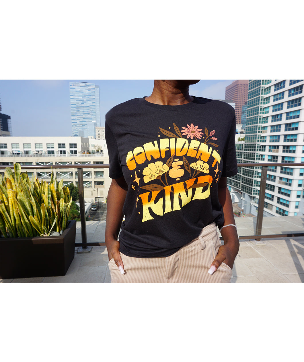 A girl modeling a blue shirt with “Confident & Kind” in half yellow and half orange organic fond. The words are surrounded by various flowers and stars - from Sierra Schultzzie