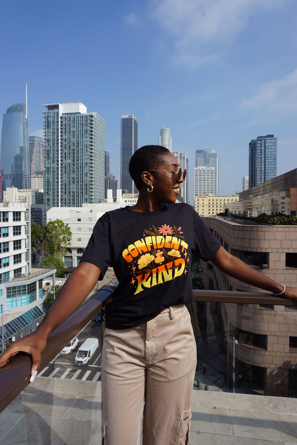 A girl modeling a blue shirt with “Confident & Kind” in half yellow and half orange organic fond. The words are surrounded by various flowers and stars - from Sierra Schultzzie