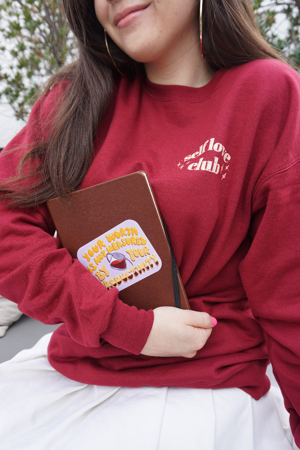 A rectangular purple sticker with curved edges on a brown notebook held by a girl. “Your worth is not measured by your productivity” is in yellow organic font with white highlights. A vector drawn coffee pot is between “by” and “your” and is filling up the word” productivity” with coffee - from Sierra Schultzzie