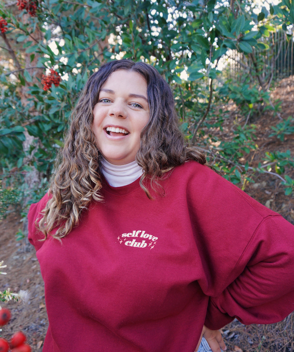 A woman modeling a red crewneck sweatshirt with “Self love” in white serif font outlined in orange arched above “club” in white serif font outline in orange. “Club” is surrounded by orange stars - from Sierra Schultzzie
