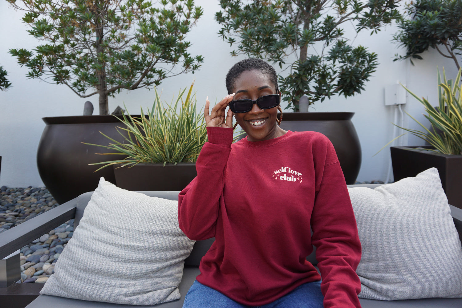 A woman modeling a red crewneck sweatshirt with “Self love” in white serif font outlined in orange arched above “club” in white serif font outline in orange. “Club” is surrounded by orange stars - from Sierra Schultzzie