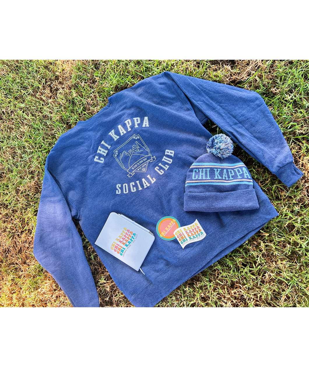 A blue crewneck that reads "Chi Kappa; Social Club" laying on grass with a white journal, two stickers and a blue knit beanie on top. All part of the Chi Kappa collection from Sierra Schultzzie.