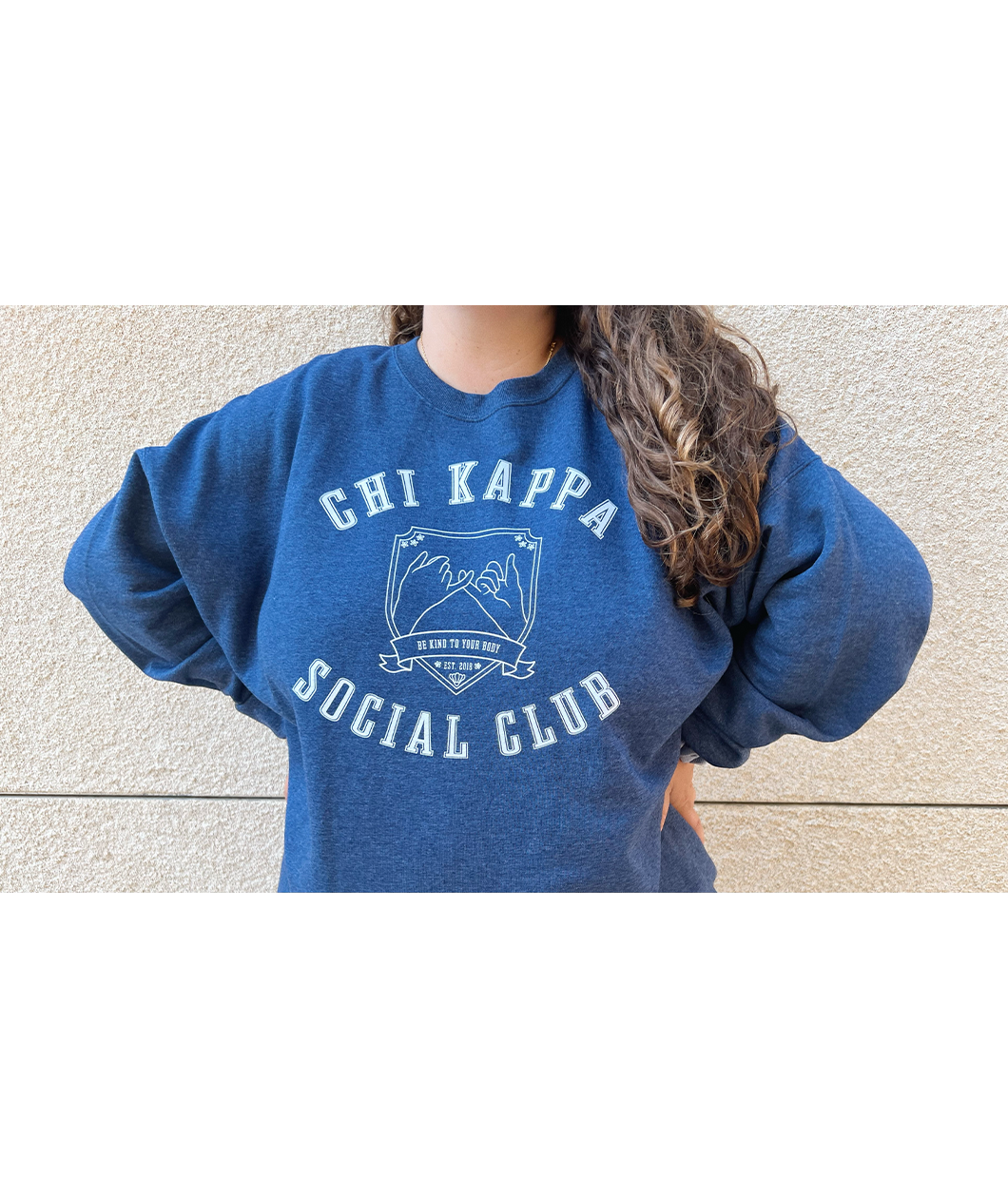 A person wearing an oversized blue crewneck that reads "Chi Kappa; Social Club" across the front with a picture of two pinkys intertwined. From Sierra Schultzzie.