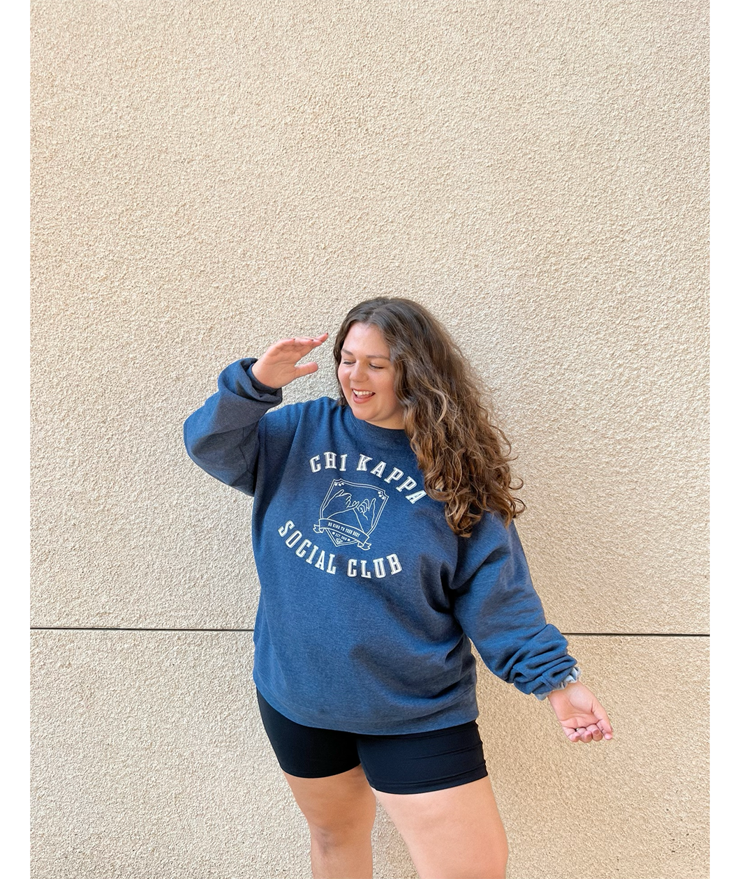 A person wearing an oversized blue crewneck that reads "Chi Kappa; Social Club" across the front with a picture of two pinkys intertwined. From Sierra Schultzzie.