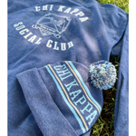 A blue knit beanie that reads "Chi Kappa" laying on top of a blue crewneck in the grass. From Sierra Schultzzie.