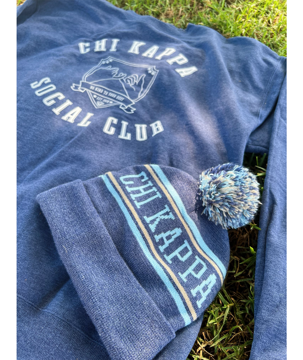 A blue knit beanie that reads "Chi Kappa" laying on top of a blue crewneck in the grass. From Sierra Schultzzie.