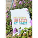 A white journal that has the words "Chi Kappa" written in four different colors on the front. Below that it says "Social Club Est. 2018". The journal is sitting among purple flowers. From Sierra Schultzzie. 