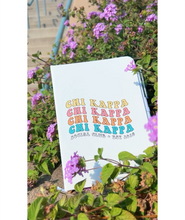 A white journal that has the words "Chi Kappa" written in four different colors on the front. Below that it says "Social Club Est. 2018". The journal is sitting among purple flowers. From Sierra Schultzzie. 