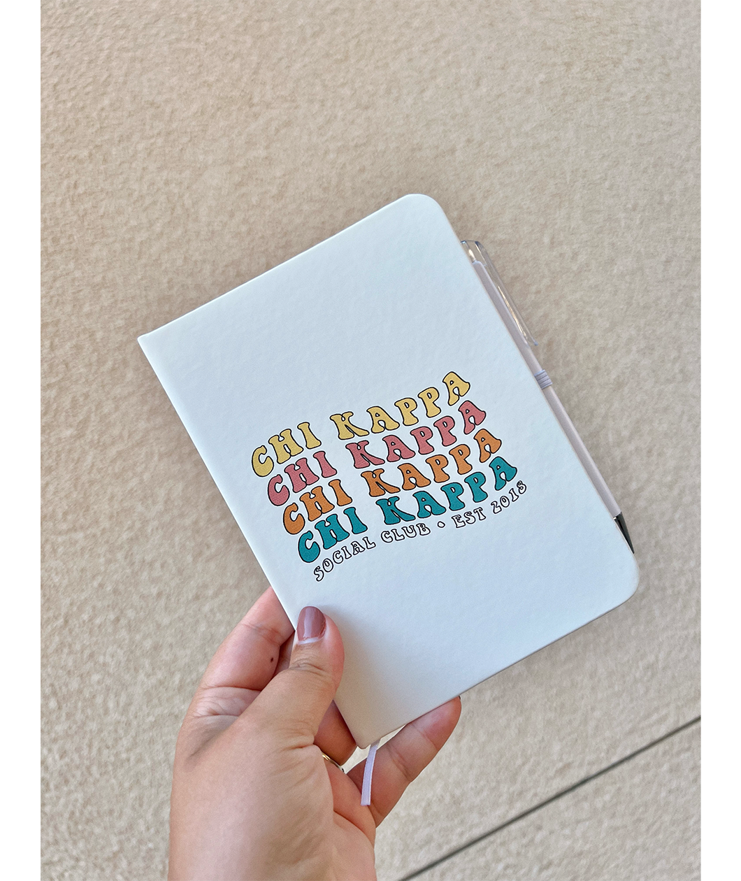 A white journal that has the words "Chi Kappa" written in four different colors on the front. Below that it says "Social Club Est. 2018". The journal is being held by a hand. From Sierra Schultzzie.