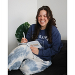A person is sitting on a couch, laughing, while writing in a white journal that says Chi Kappa. From Sierra Schultzzie. 