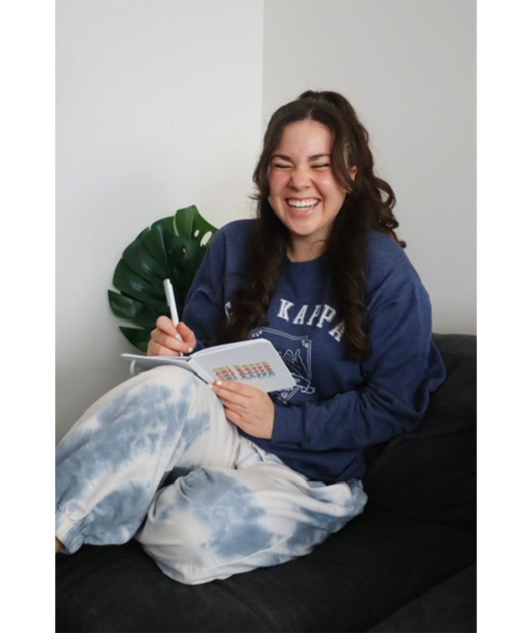 A person is sitting on a couch, laughing, while writing in a white journal that says Chi Kappa. From Sierra Schultzzie. 