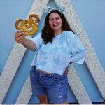 Sierra Schultzzie wearing jean shorts, the blue tie-dye confident and kind crop top, mickey mouse ears and holding a mickey mouse pretzel. 