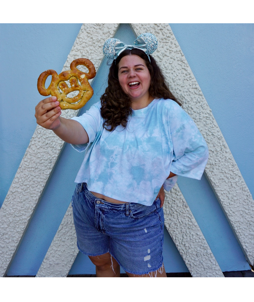 Sierra Schultzzie wearing jean shorts, the blue tie-dye confident and kind crop top, mickey mouse ears and holding a mickey mouse pretzel. 