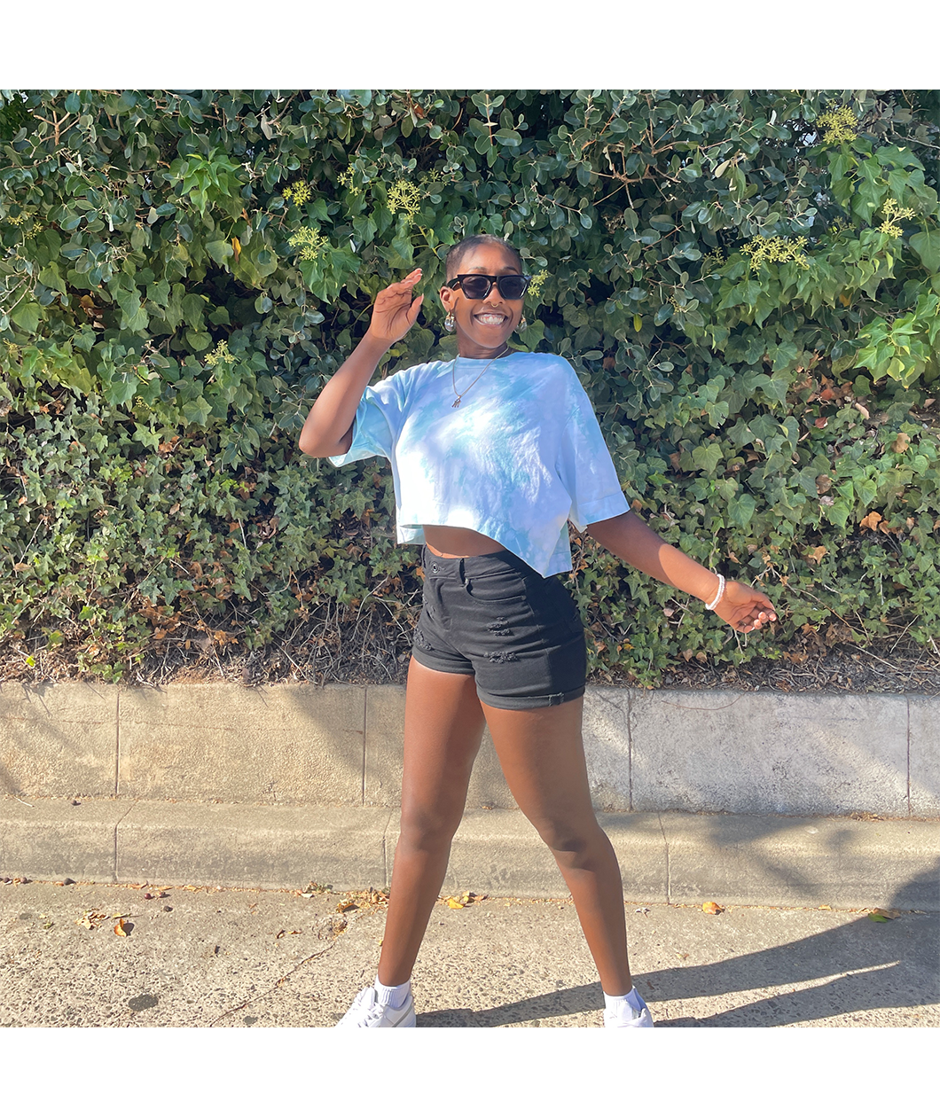 Someone wearing the blue confident and kind crop top from Sierra Schultzzie, jean shorts and sunglasses, and facing the camera smiling.