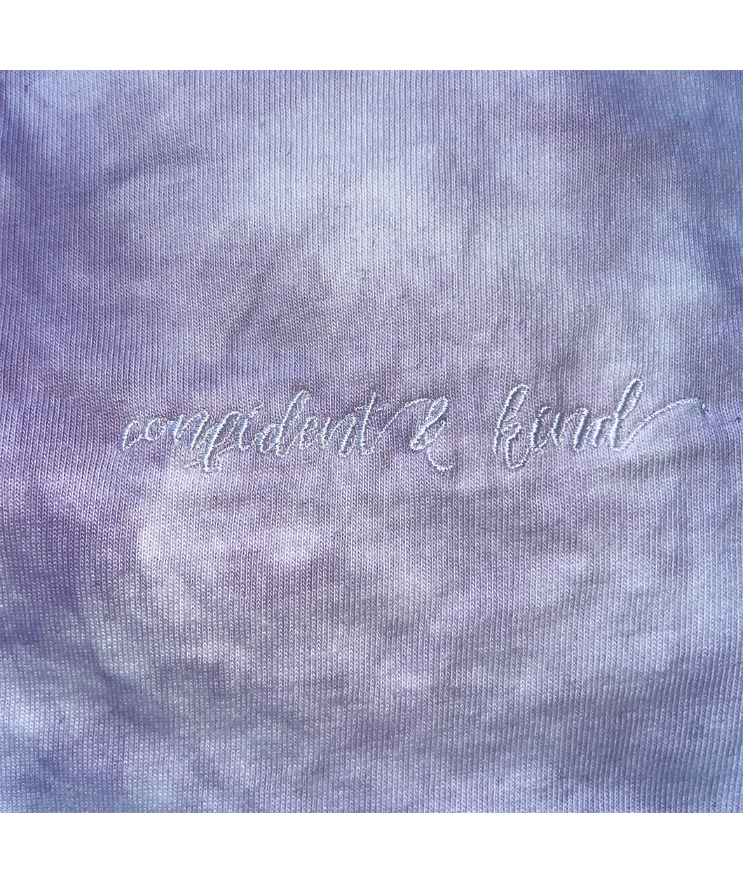 A close up of the lavender tie-dye crop top from Sierra Schultzzie, showing the embroidered words "confident & kind".