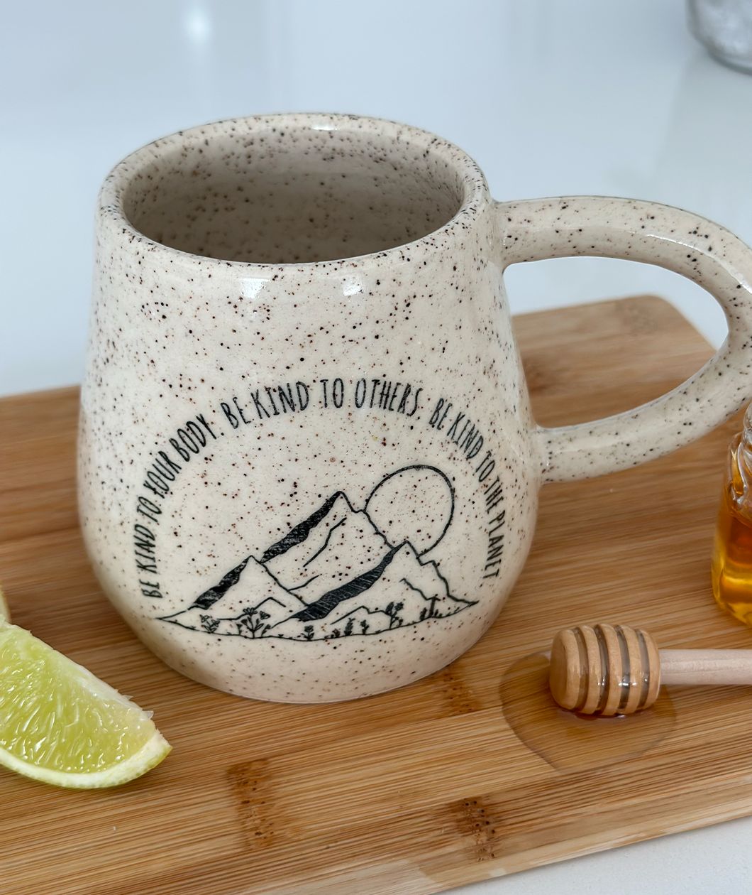 A close up of the Be Kind mug from Sierra Schultzzie on a cutting board with a honey wand next to it. The mug is speckled beige with mountains and a moon. The text curves around the mountains and says "Be kind to your body be kind to others be kind to the planet".