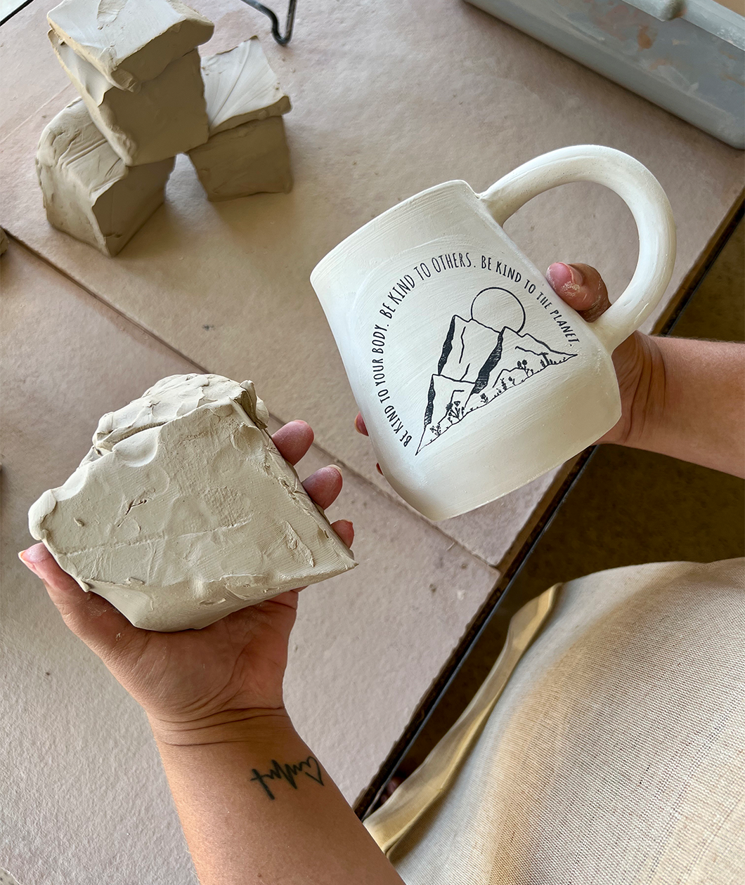 The Be Kind mug from Sierra Schultzzie in the unfinished form with a lump of clay next to it. The mug is beige with mountains and a moon. The text curves around the mountains and says "Be kind to your body be kind to others be kind to the planet".