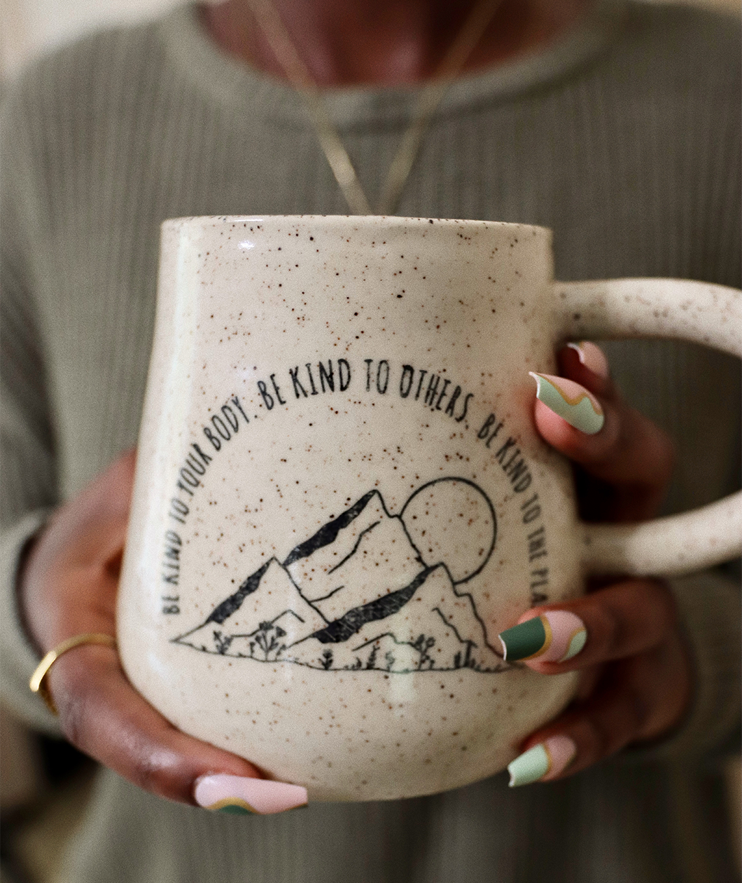 A close up of the Be Kind mug from Sierra Schultzzie in someones hands. The mug is speckled beige with mountains and a moon. The text curves around the mountains and says "Be kind to your body be kind to others be kind to the planet".