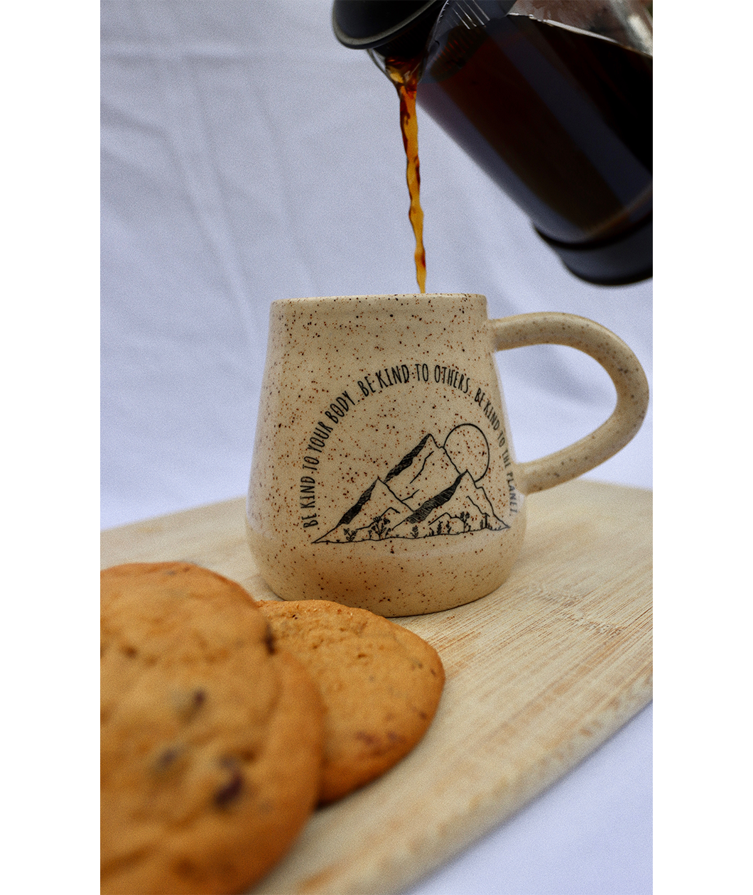 A close up of the Be Kind mug from Sierra Schultzzie on a cutting board with cookies a a french press pouring coffee in the mug. The mug is speckled beige with mountains and a moon. The text curves around the mountains and says "Be kind to your body be kind to others be kind to the planet".