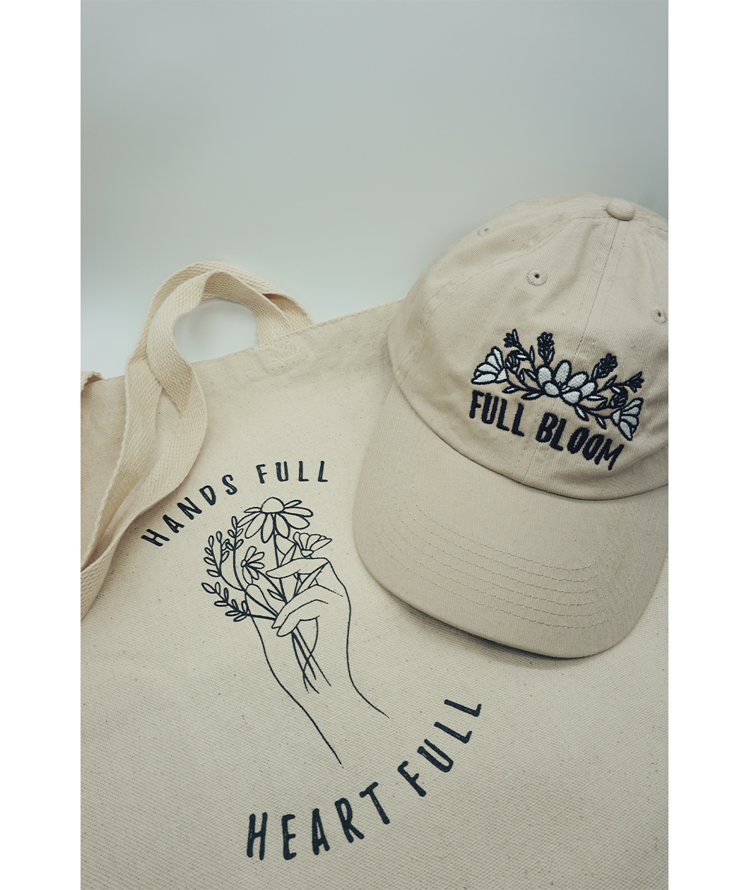 Close up of the canvas tote from Sierra Schultzzie with the words "Hands Full, Heart Full" printed in black as well as an outline of a hand holding flowers. On top of the tote in a beige ball cap with the embroidered words "Full Bloom" and flowers.