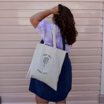 Sierra Schultzzie facing away from the camera, holding her hair with a Hands Full, Heart Full canvas tote over her shoulder. 
