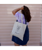 Sierra Schultzzie facing away from the camera, holding her hair with a Hands Full, Heart Full canvas tote over her shoulder. 