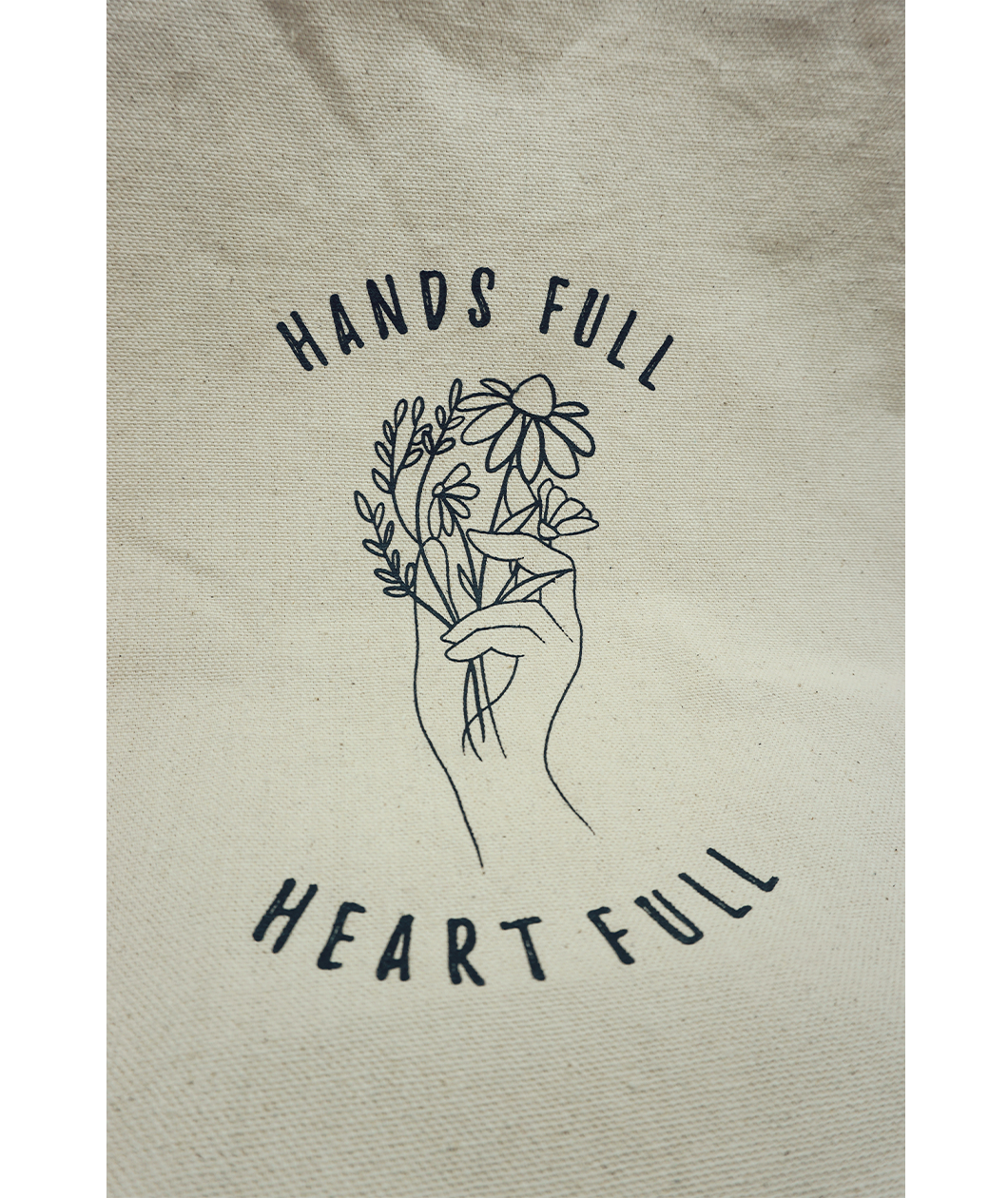 Close up of the canvas tote from Sierra Schultzzie with the words "Hands Full, Heart Full" printed in black as well as an outline of a hand holding flowers. 