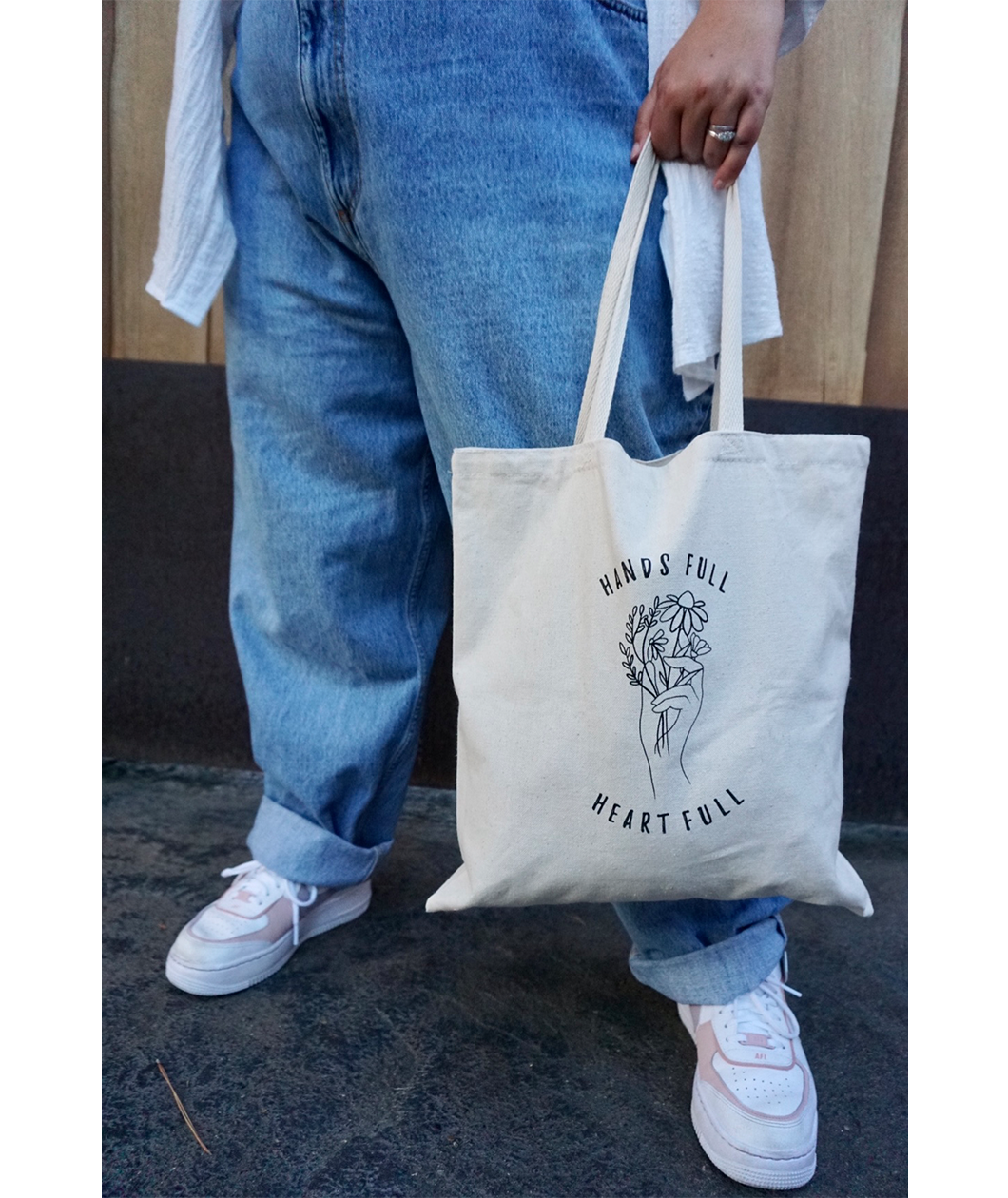 Close up of someone holding the canvas tote from Sierra Schultzzie with the words "Hands Full, Heart Full" printed in black as well as an outline of a hand holding flowers.