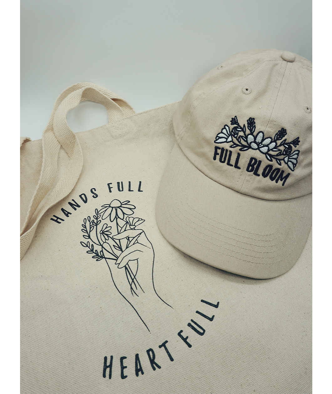 Close up of the canvas tote from Sierra Schultzzie with the words "Hands Full, Heart Full" printed in black as well as an outline of a hand holding flowers. On top of the tote in a beige ball cap with the embroidered words "Full Bloom" and flowers. 