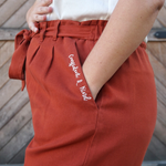 A woman modeling copper colored pants with a copper colored belt tied in a knot. “Confident & Kind” is in white curisve font along the lefthand pocket - from Sierra Schultzzie
