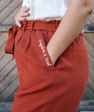 A woman modeling copper colored pants with a copper colored belt tied in a knot. “Confident & Kind” is in white curisve font along the lefthand pocket - from Sierra Schultzzie
