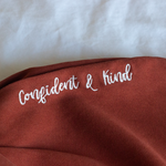 Confident & Kind” is in white curisve font along the lefthand pocket of copper colored pants - from Sierra Schultzzie