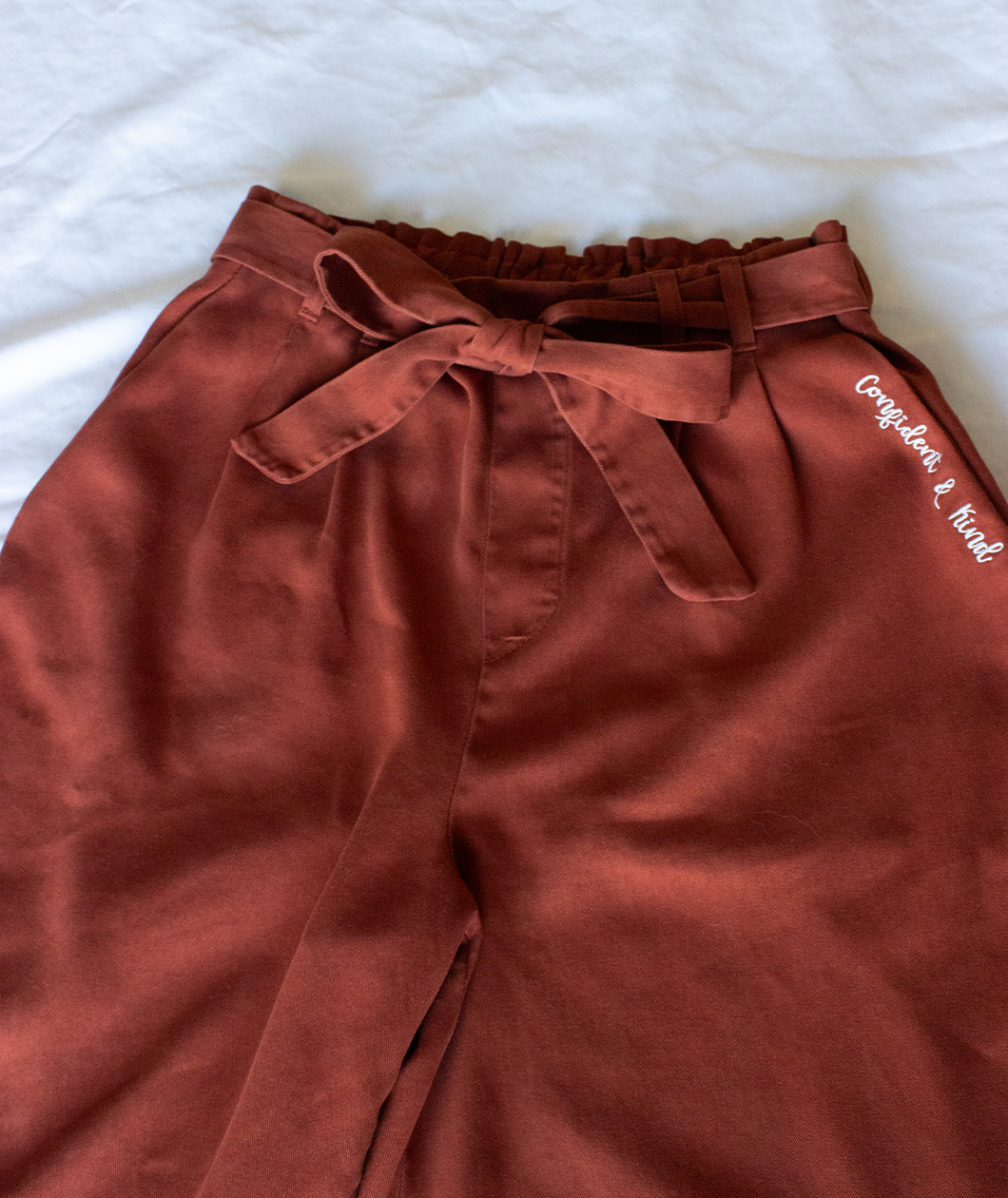 Copper colored pants with a copper colored belt tied in a knot. “Confident & Kind” is in white curisve font along the lefthand pocket - from Sierra Schultzzie