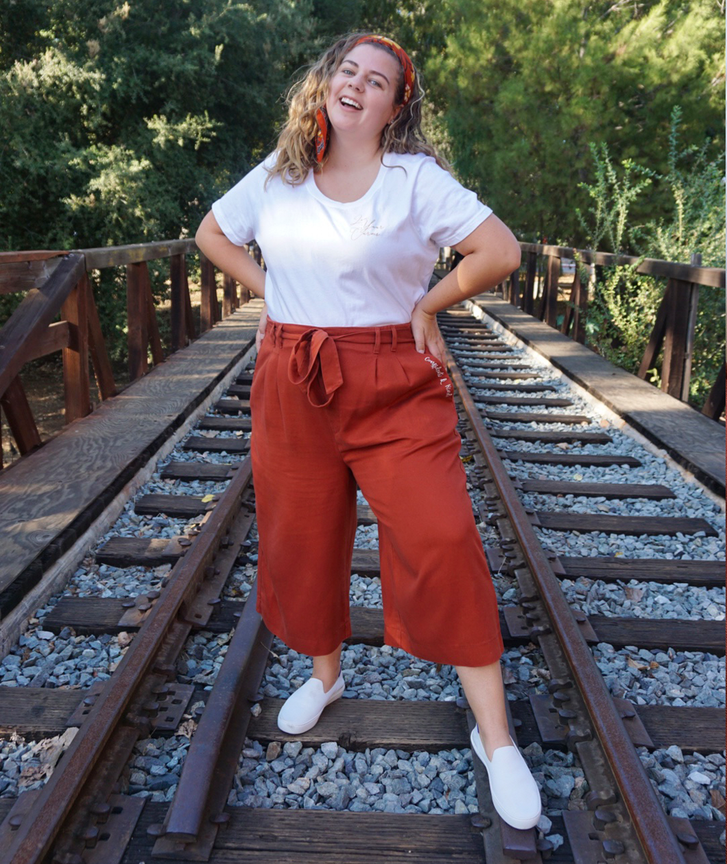 A woman modeling copper colored pants with a copper colored belt tied in a knot. “Confident & Kind” is in white curisve font along the lefthand pocket - from Sierra Schultzzie