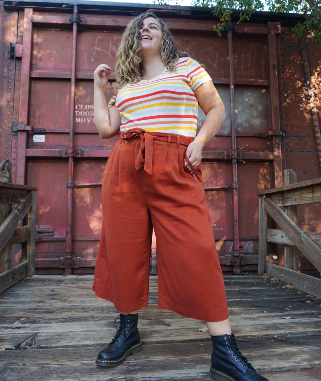 A woman modeling copper colored pants with a copper colored belt tied in a knot. “Confident & Kind” is in white curisve font along the lefthand pocket - from Sierra Schultzzie