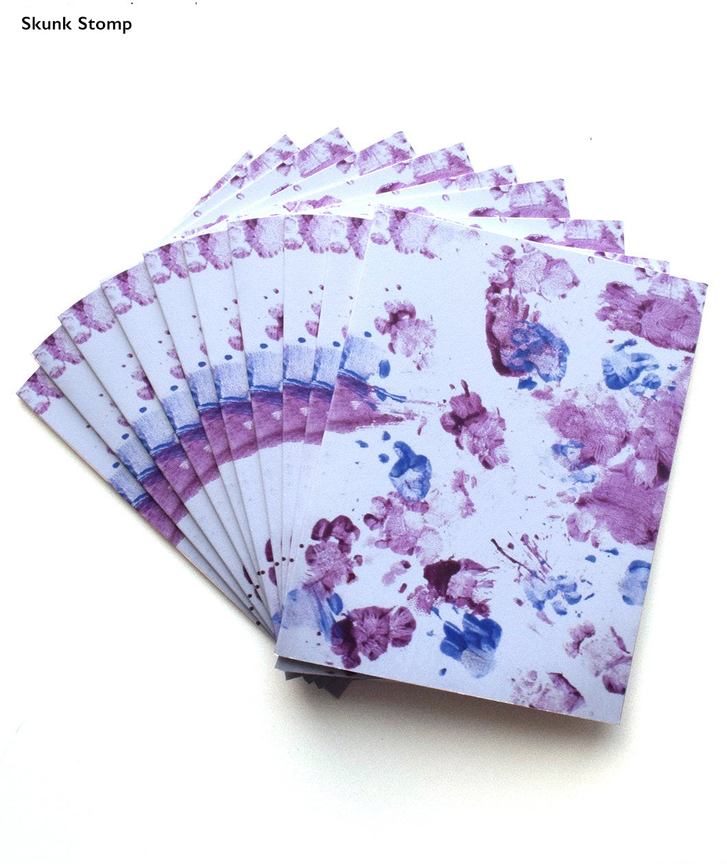Purple and blue footprints made by skunk and cavy on greeting cards. Multple greeting cards are fanned out from behind. “Skunk Stomp” is written in top left of image in black sans serif font indicating which greeting card is being shown - from Animal Wonders