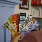 Someone holding a fan of 8 different illustrated, colorful bookmarks from the Books Unbound subscription. 