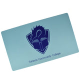 A rectangular mousepad-like mat in light blue. Centered on the mat is the TCC logo – a dragon's head peering at the pages of an open book, with a crest shape in the background. The blue text “Tolarian Community College” is below the logo.