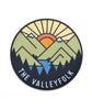 A circular sticker with a vector drawing of a mountain scene, and trees with a sun rising in the background. "The Valleyfolk" is arched at the bottom in light blue sans serif font - from the Valleyfolk