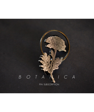 A gold pin shaped like a flower with the text "Botanica; Pin Subscription".