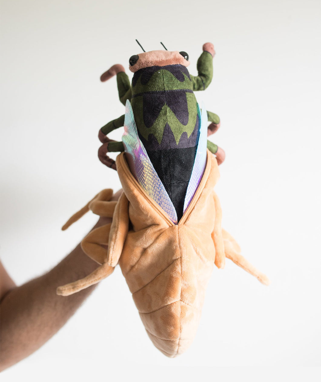 A green, black, and grey cicada held aloft by Tyler Thrasher. The cicada has iridescent wings and is halfway emerged from its tan plush shell, which is unzipped.