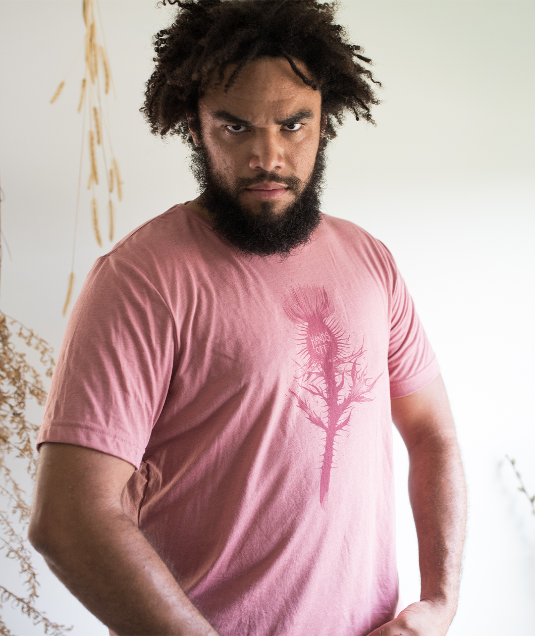 Tyler Thrasher modeling a red shirt with a dark red silhouette of a thistle weed in the center of the shirt. “Hands off” is written on the top of the thistle weed in red sans serif font - from Tyler Thrasher