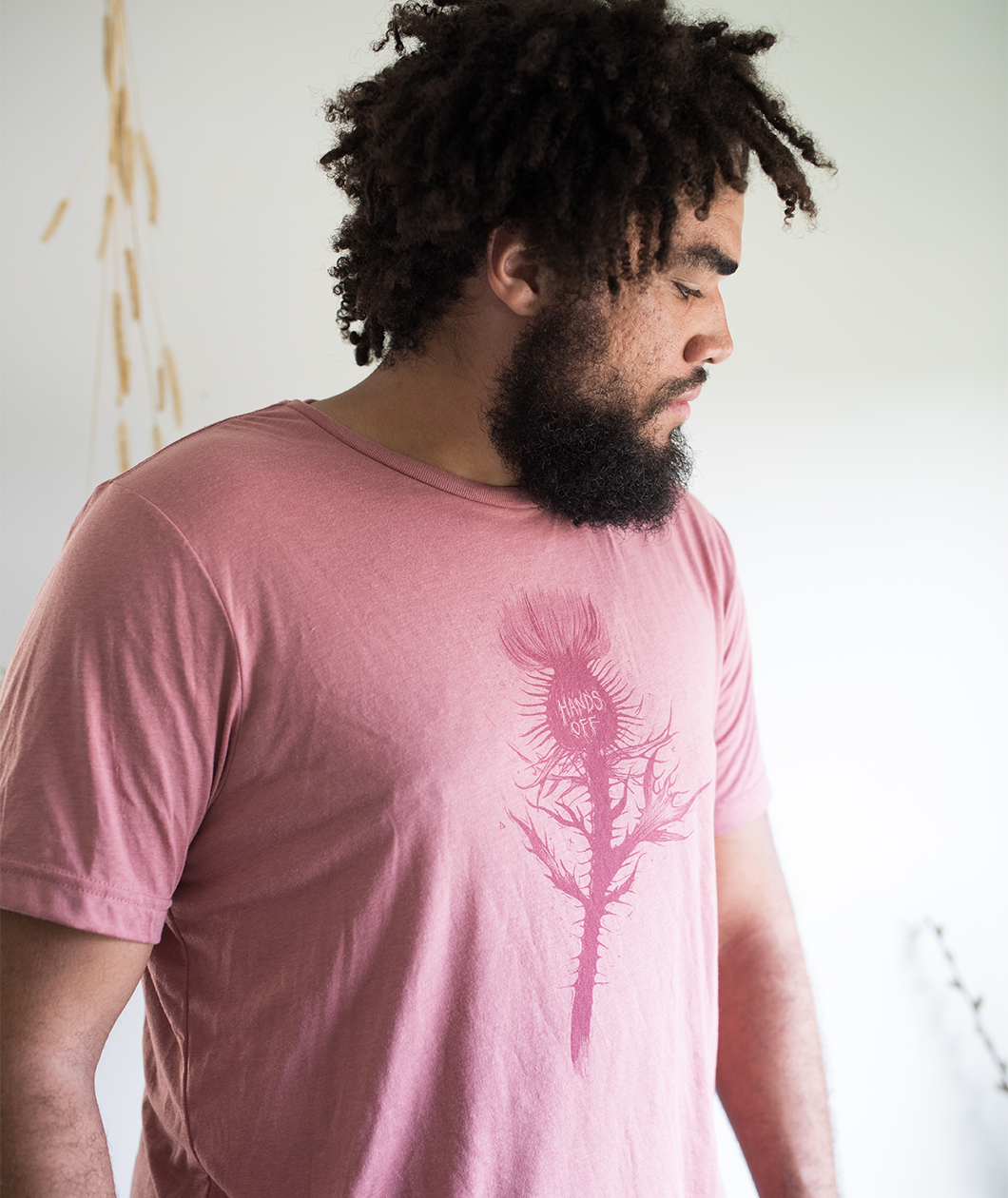Tyler Thrasher modeling a red shirt with a dark red silhouette of a thistle weed in the center of the shirt. “Hands off” is written on the top of the thistle weed in red sans serif font - from Tyler Thrasher