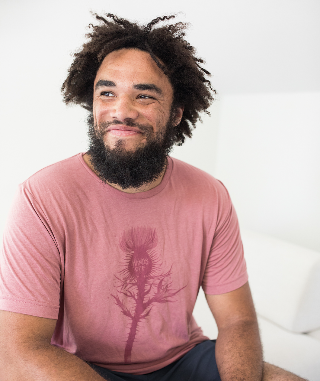 Tyler Thrasher modeling a red shirt with a dark red silhouette of a thistle weed in the center of the shirt. “Hands off” is written on the top of the thistle weed in red sans serif font - from Tyler Thrasher